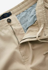 Beige színű pamut sort elasztikus derékkal, cipzárral és gombos záródással, kék és fehér csíkos bélés jellemzi.