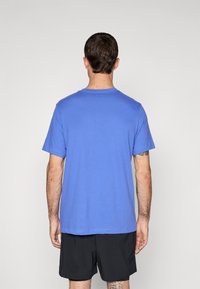 Hombre con cabello corto y oscuro vistiendo una camiseta de manga corta azul lisa y pantalones cortos negros, visto desde atrás contra un fondo liso.