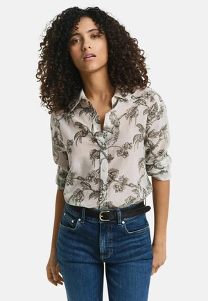 Chemise à boutons en tissu léger avec un imprimé floral et d'oiseaux, dotée d'un col évasé et de manches retroussées, associée à un jean foncé et une ceinture noire.