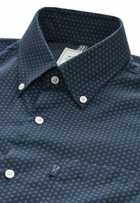 Chemise pour homme bleu foncé à boutons avec un petit motif floral bleu clair et des boutons blancs, montrant le col et la zone de la poche avant.