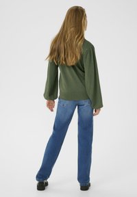 Pull en tricot vert avec des manches texturées, associé à un jean droit bleu. Le modèle se tourne, mettant en valeur l'arrière de la tenue.