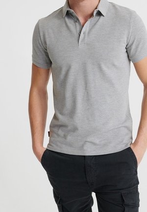 Homme portant un polo gris clair à manches courtes et un pantalon cargo noir, les mains dans les poches, sur un fond uni.