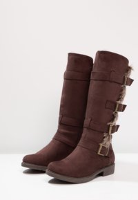 Botas de ante marrón hasta la mitad de la pantorrilla con tres correas laterales y hebillas, ribete de piel sintética en el lado exterior y tacones bajos apilados.