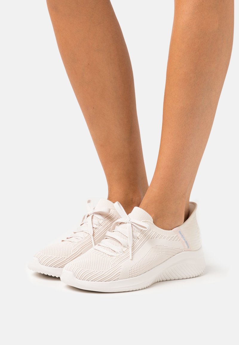 Jambes portant des baskets tricotées beige clair avec lacets et semelles blanches côtelées sur fond blanc uni.
