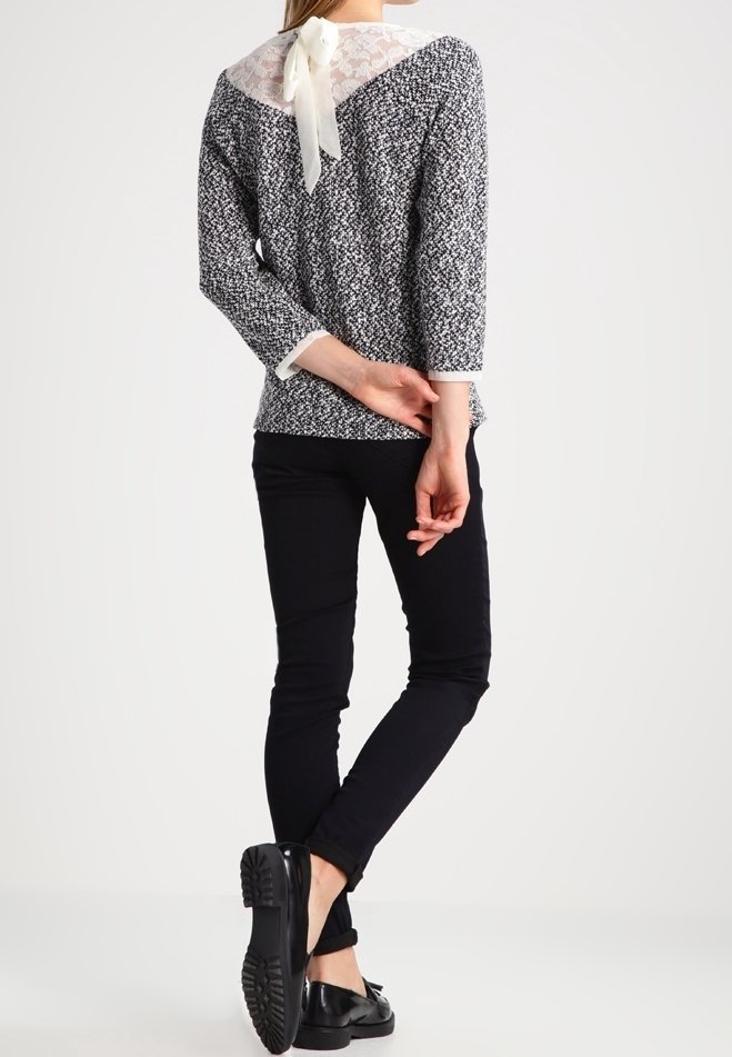 Svart och vit mönstrad blus med spetsbak och rosett vid halsringningen; bärs med åtsittande svarta byxor och svarta skor.