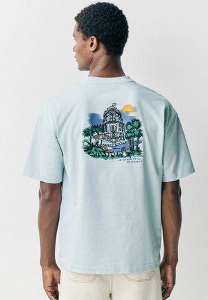 Camiseta de algodón de color azul claro con mangas cortas, que presenta una impresión gráfica de un hotel y palmeras en la parte posterior. Diseño simple, textura suave.