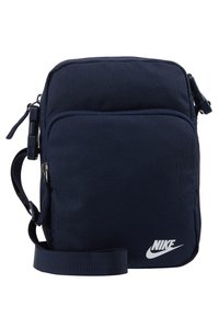 Tmavě modrá látková kabelka přes rameno s nastavitelným popruhem, přední zipová kapsa a bílé logo Nike. Obdélníkový tvar s texturovaným povrchem.