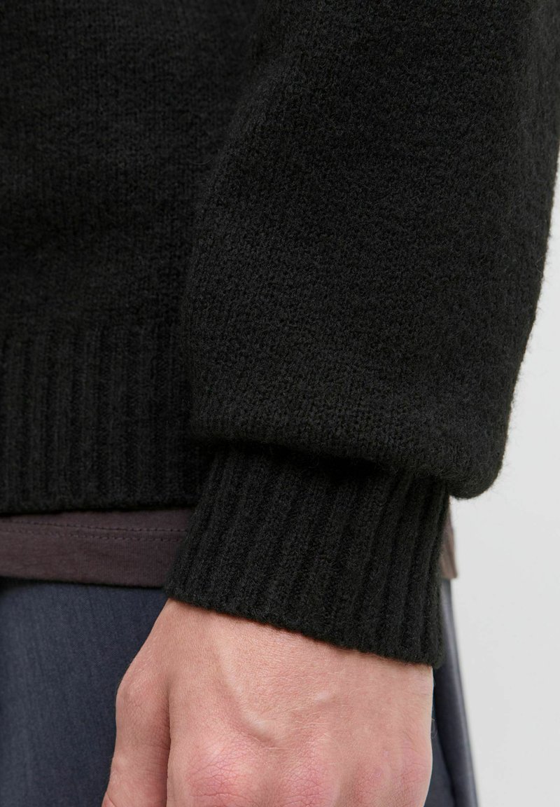 Gros plan sur la main et le poignet d'une personne portant une manche de pull noir en maille côtelée, avec une chemise marron foncé et un pantalon bleu marine visibles.