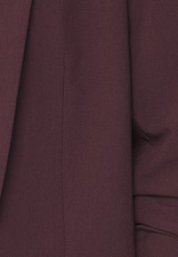 Lähikuva syvän violetista villatakin hihasta, jossa näkyy kankaan rakenne ja käsityönä tehdyt ompeleet hihan ja sivusauman varrella.