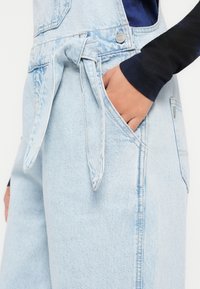 Lichtblauwe denim tuinbroeken met een strikdetail in de taille, zijzak en vervaagde textuur, voorzien van zilveren hardware en contrasterende stiksels.