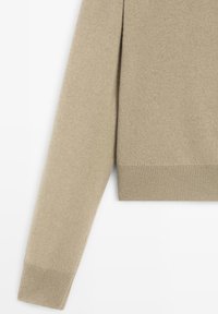 Beige lange mouw gebreide trui, gedeeltelijk weergegeven op een witte achtergrond, met geribbelde manchetten en zoom.