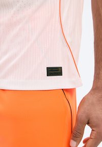 Κοντινό πλάνο ενός ατόμου που φορεί ροζ ανοιχτού χρώματος αθλητική μπλούζα με τρύπες και ετικέτα, μαζί με φωτεινά πορτοκαλί σορτς με μαύρες ραφές.