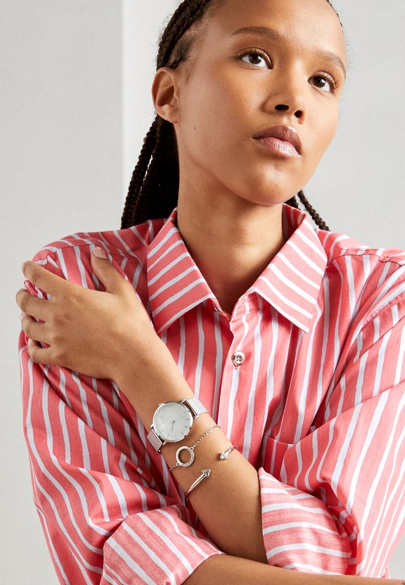Silver analog watch with a mesh band, worn on a wrist adorned with three bracelets. The model wears a pink striped button-up shirt.