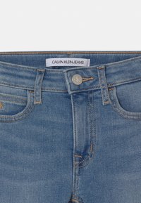 Acercamiento de la cinturilla de unos pantalones vaqueros azul de Calvin Klein, con un botón de metal que presenta el logo de CK y con lazos para cinturón visibles.