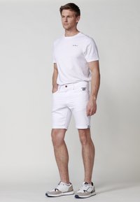 Camiseta blanca de cuello redondo con un pequeño logotipo, combinada con pantalones cortos blancos arremangados que cuentan con un cierre de botón y una marca bordada. Zapatillas blancas.