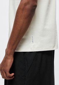 Jasnobeżowy t-shirt z bawełny, z krótkimi rękawami i okrągłym dekoltem, z bocznym rozcięciem i widoczną etykietą marki u dołu. Widoczne czarne spodnie.