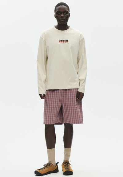 Jeune homme portant une chemise beige à manches longues avec un logo, un short orange à carreaux, des chaussettes beiges et des chaussures orange à lacets noirs, debout sur un fond blanc.