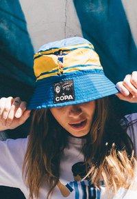 Blauw en geel emmerhoed van een gestructureerde stof, met een centraal logo-patch en borduurdetails bovenop.