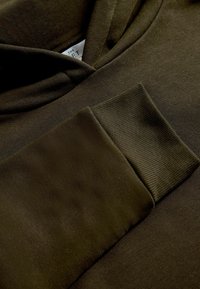 Dunkelgrüner Kapuzenpullover mit weicher Textur, gerippten Bündchen und lässigem Design. Enthält ein Etikett mit Markeninformationen.