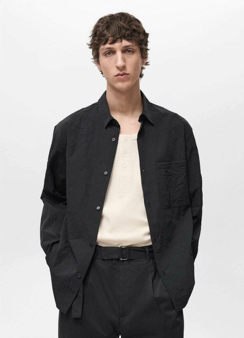 Schwarze, zerknitterte Knopfhemd mit einer einzelnen Tasche, darüber getragen ein hellbeiges Henley. Kombiniert mit schwarzen Hosen und einem Gürtel.