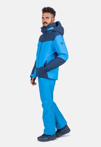 Blaue wasserdichte Jacke mit Kapuze, die dunklere blaue Akzente aufweist, kombiniert mit blauen wasserdichten Hosen und robusten schwarzen Stiefeln.