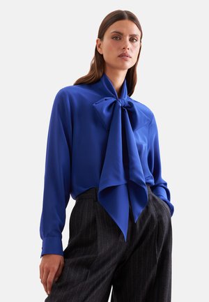 Femme portant un chemisier bleu cobalt à manches longues avec un grand nœud noué au cou et un pantalon rayé foncé, debout avec une main dans la poche.
