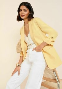 Kobieta w żółtej marynarce, białej koronkowej bluzce i białych spodniach, opierająca się na drewnianym krześle z plecionym siedziskiem na beżowym tle.