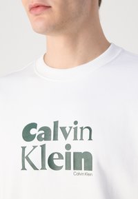 Biela mikina s rebrovaným okrúhlym výstrihom, s veľkým zeleným grafickým nápisom "Calvin Klein" v rôznych písmach na hrudi.
