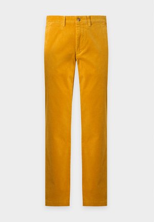 Homme debout portant une chemise à carreaux vert foncé et violet à boutons, un pantalon jaune moutarde et des baskets blanches sur un fond uni.