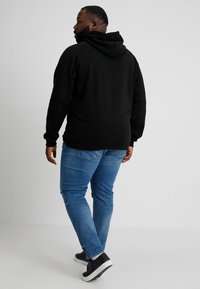 Felpa nera con una vestibilità comoda, abbinata a jeans di denim blu e sneakers nere. Il tessuto sembra morbido, con una tasca a marsupio ben visibile.