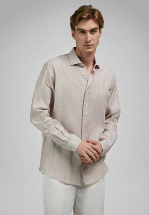 Jeune homme portant une chemise à manches longues beige à fines rayures, avec un pantalon blanc, debout les mains croisées devant un fond gris.