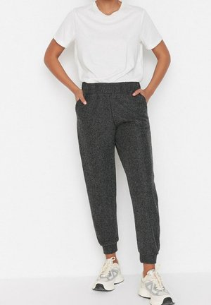 Person iført en hvid t-shirt med korte ærmer, mørkegrå strikkede joggebukser og hvide sneakers, stående med hænderne i lommerne.