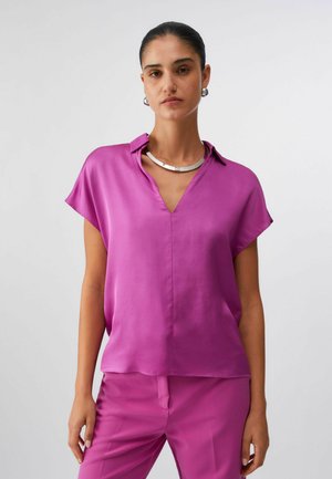 Donna con camicetta a maniche corte di un viola acceso e pantaloni coordinati, accessoriate con collana d'argento e orecchini a cerchio, in piedi davanti a uno sfondo bianco.