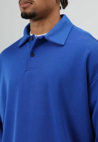 Blauw poloshirt gemaakt van zacht materiaal. Voorzien van een kraag, een knoopsluiting met drie knopen en een ontspannen pasvorm. Gladde textuur met minimale details.