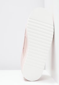Semelle d'une basket rose clair avec une épaisse semelle extérieure en caoutchouc blanc à rainures, inclinée contre des surfaces blanches.