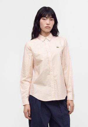 Femme aux longs cheveux foncés portant une chemise rose pâle boutonnée et un pantalon bleu marine, debout devant un fond clair uni.