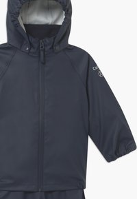Marineblauwe waterdichte jas met capuchon, ritssluiting, elastische manchetten en een gladde textuur. Heeft logo-opdruk op de mouw.