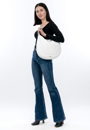 Weiße Leder-Umhängetasche mit geschwungenem Design, glatter Textur und subtiler Markenkennzeichnung am Boden. Getragen von einem Model, kombiniert mit ausgestellten Jeans.