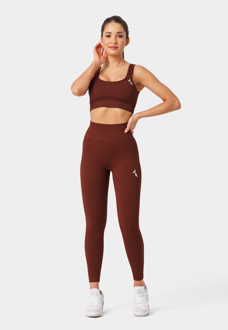 Prążkowany sportowy top i legginsy w ciemnym brązie, wykonane z elastycznego materiału. Posiadają wysoki stan i biały akcent z logo. Sneakersy w zestawie.