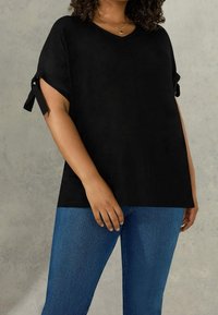 Black V-neck top with short sleeves featuring tie accents. Smooth texture, relaxed fit, paired with light blue fitted jeans.