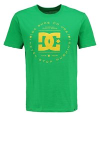 Zelené bavlněné tričko s žlutým grafickým potiskem, na kterém je logo "DC", hvězdičky a text "NEVER STOP PUSHING" uspořádaný v kruhu.