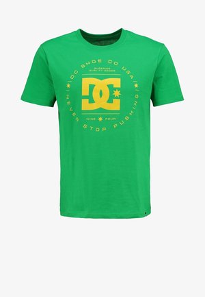 T-shirt en coton vert avec un logo jaune, pantalon ajusté gris clair et chaussures noires. La personne tient un sac à dos foncé avec des accents rouges.
