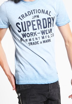 Person iført en lyseblå t-shirt med mørkeblå "Superdry Work-Wear" tekst og sorte bukser med lynlåslomme.