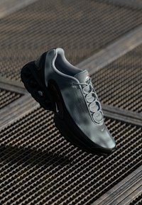 Sive Nike tenisice z mrežasto zgornjo plastjo, ukrivljenim črnim podplatom z zračnimi blazinicami in poenostavljenim dizajnom vezalk.
