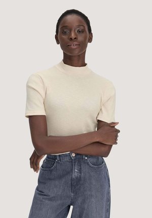 Femme à la peau foncée portant un haut côtelé crème à manches courtes et un jean bleu taille haute, debout les bras croisés.