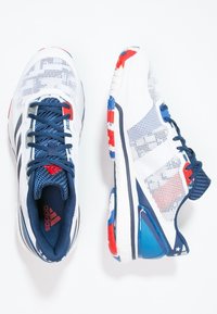 Scarpe da ginnastica bianche con tomaia in rete, lacci blu navy, accenti rossi e blu, e una suola strutturata con un tallone imbottito.