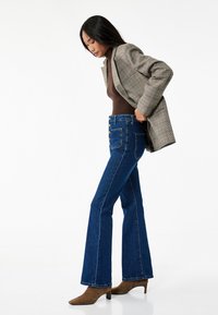 Uppvikta blå denimjeans med klassisk femfickdesign, knäppning framtill och kontrasterande sömmar, ihopparade med en brun figurnära topp och en rutig blazer.