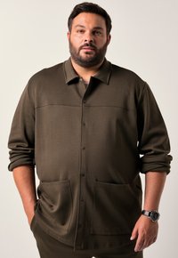 Muž se světlým plnovousem, oblečený v olivově zelené bundě na knoflíky s velkými předními kapsami a vyhrnutými rukávy, stojí s rukama v kapsách.