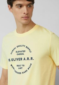 Geel katoenen T-shirt met een cirkelvormig zwart grafisch ontwerp met tekst. Korte mouwen, ronde hals en een zachte textuur.