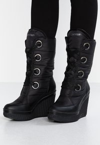 Botines negros de cuña con exterior brillante de nailon, con cordones delanteros, ojales metálicos y suela de cuña texturizada. Logo de la marca visible.